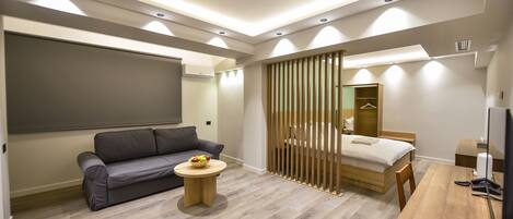 A modern hotel room with a sofa, a small round table with a fruit bowl, a wooden bed frame, a wooden desk, and a TV.