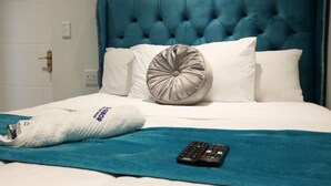 A hotel room with a tufted headboard, white bedding, a grey pillow, a white towel, and a remote control on a teal blanket. - Elgibor Best Guest House  (Cape Town)