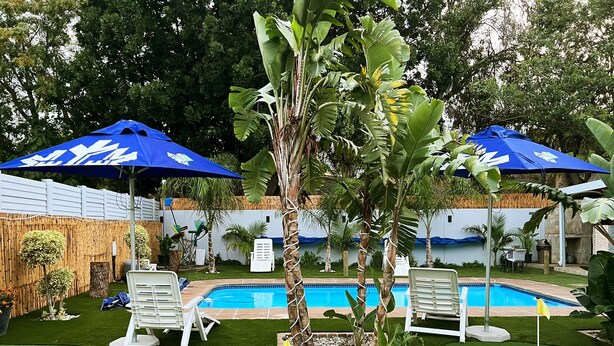 A pool area with artificial grass, two white lounge chairs, and two blue umbrellas. - Elgibor Best Guest House  (Cape Town)