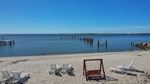 Ein Strand mit mehreren Liegestühlen und einem Schaukelstuhl, Holzpfählen im Wasser und einem klaren, blauen Himmel.
