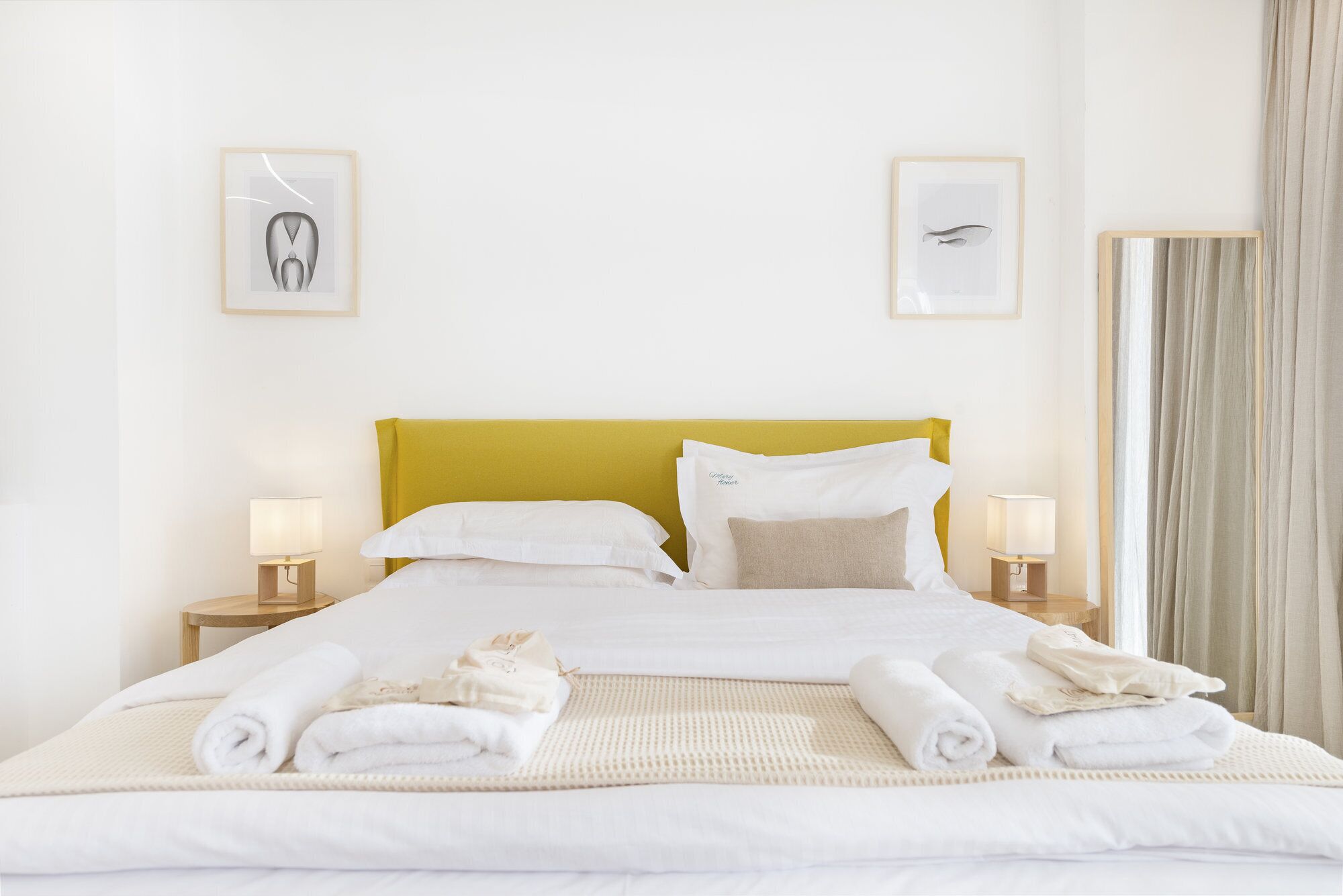 A neatly made bed with white linens and rolled towels, a mustard-colored headboard, and two bedside lamps.