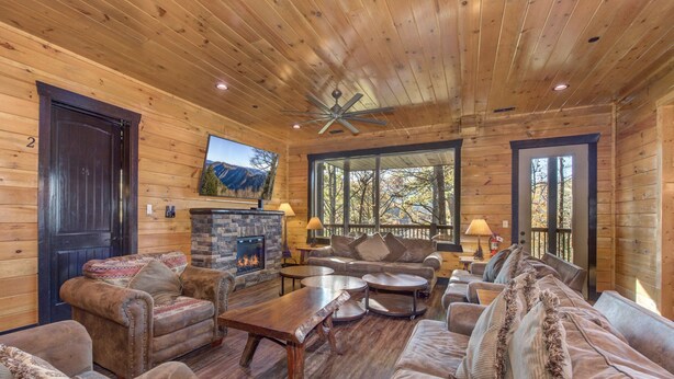 A log cabin interior with a fireplace, leather sofas, a wooden coffee table, and a large window offering a view of trees.
