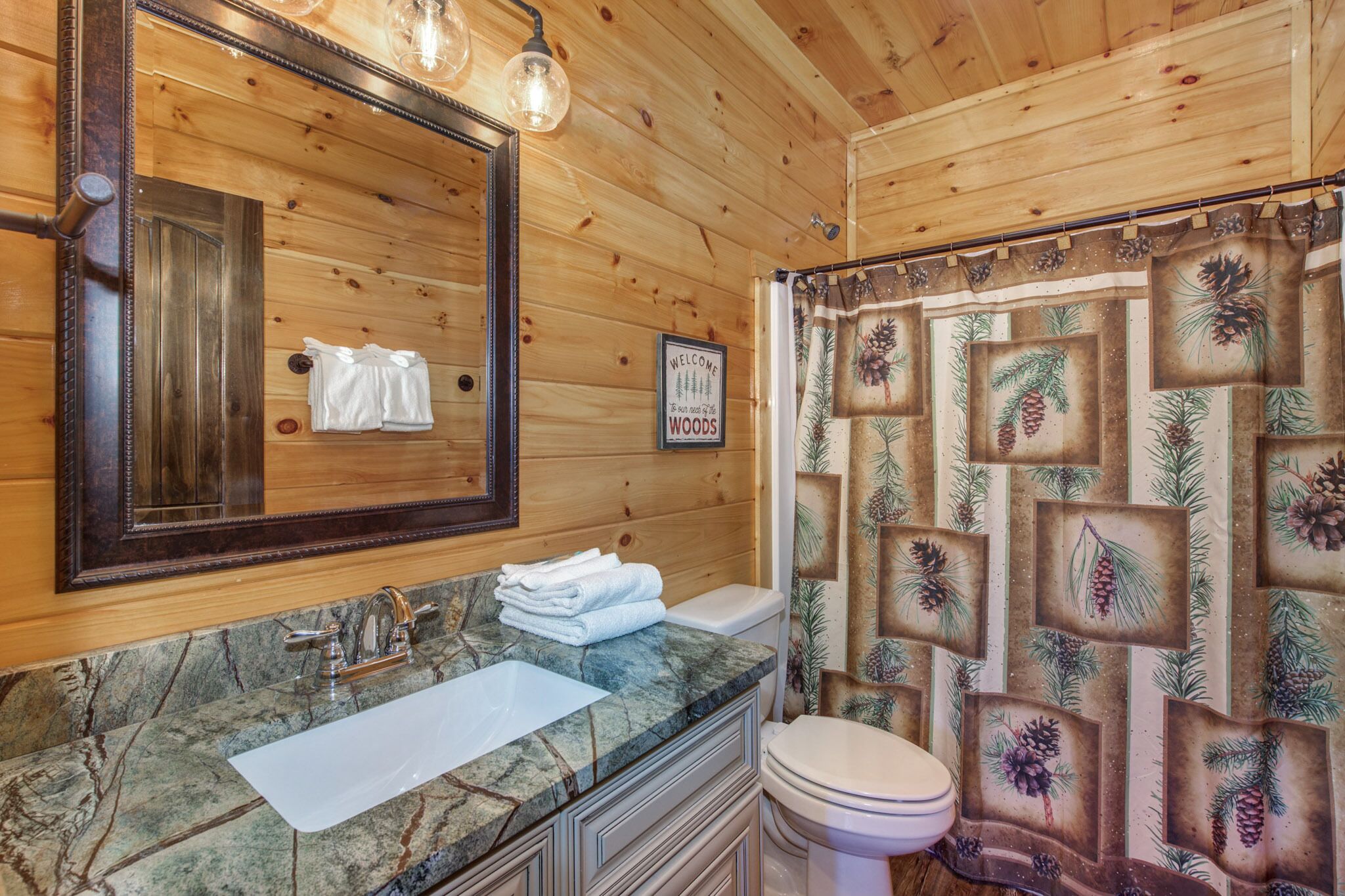Une salle de bain rustique, avec des murs en bois, un lavabo en marbre, des toilettes et un rideau de douche orné de motifs représentant des pommes de pin et des branches de pin.