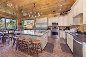 A spacious kitchen with wooden cabinetry, a large island with stools, stainless steel appliances, and a view through the windows. - Luxury Cabin Sleeps 64, Walk to Gatlinburg, Pool Access, Hot Tub (Gatlinburg)