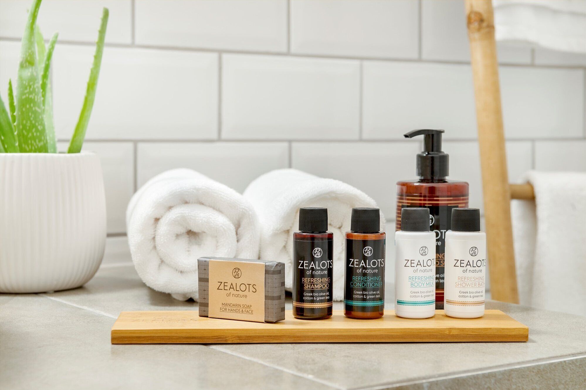 A bathroom with a wooden shelf displaying various Zealots of nature products, a rolled white towel, a bamboo brush, and a potted aloe plant.