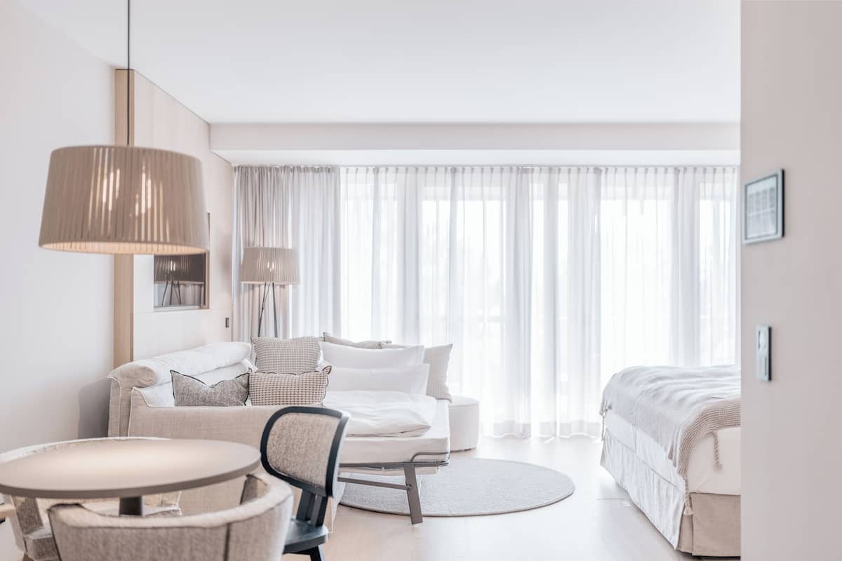 Una habitación de hotel moderna con sofá, una mesa redonda, una silla, una cama y un gran ventanal con cortinas translúcidas.