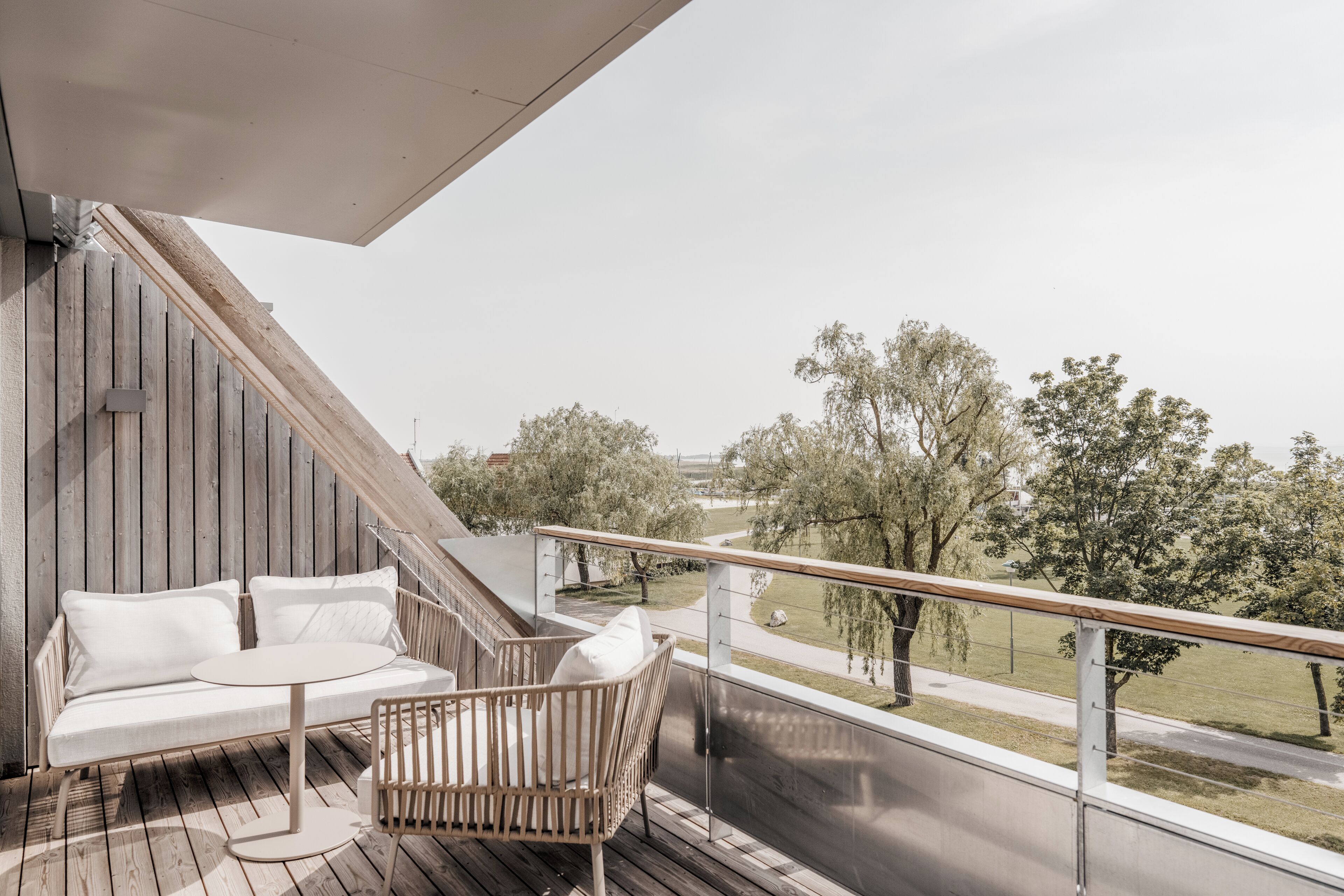A balcony with a wooden deck, a white sofa, and a wicker chair, overlooking a grassy area with trees and a road.