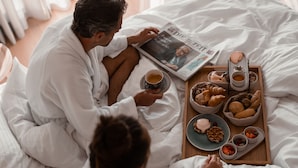 A couple in a large, well-lit bedroom with a wooden floor, sitting on a bed with a tray of breakfast items including pastries, fruits, and beverages.