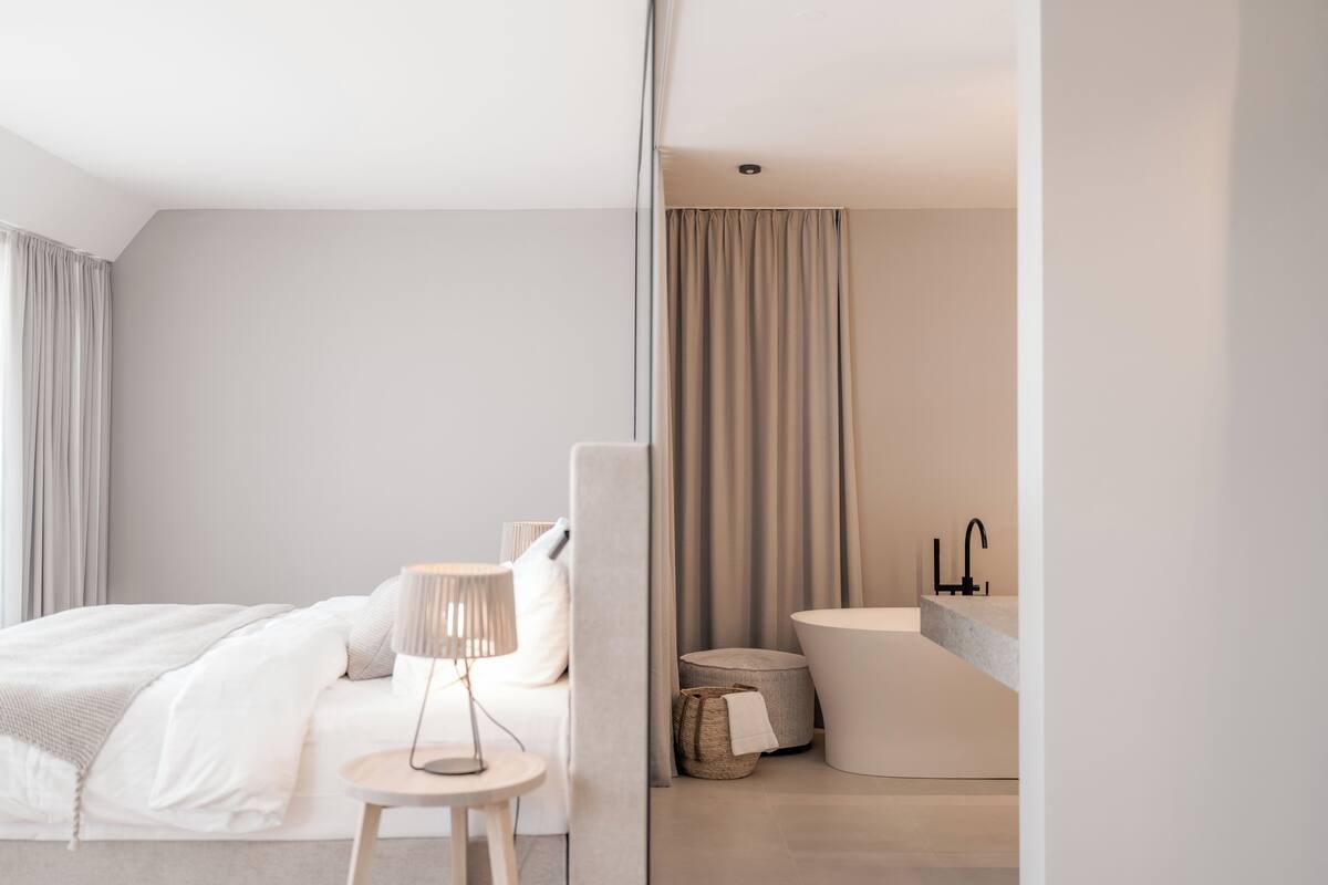 Habitación de hotel moderna con una cama grande, mesita de noche, lámpara y baño con bañera y lavamanos.