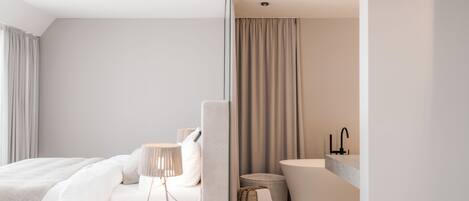 Habitación de hotel moderna con una cama grande, mesita de noche, lámpara y baño con bañera y lavamanos.