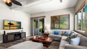 A modern living room with a sofa, ottoman, coffee table, and a painting of a surfer. - 🏖️Kulalani Townhome w/ Mountain Views + Beach Club Access (Kamuela)