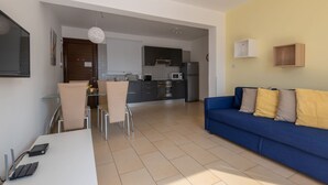 A modern kitchen with dark cabinets, a microwave, and a refrigerator. There is a dining area with a white table and chairs. A blue sofa with cushions is in the living area. - Narcissos Nissi Beach Apartment C3 (Ayia Napa)
