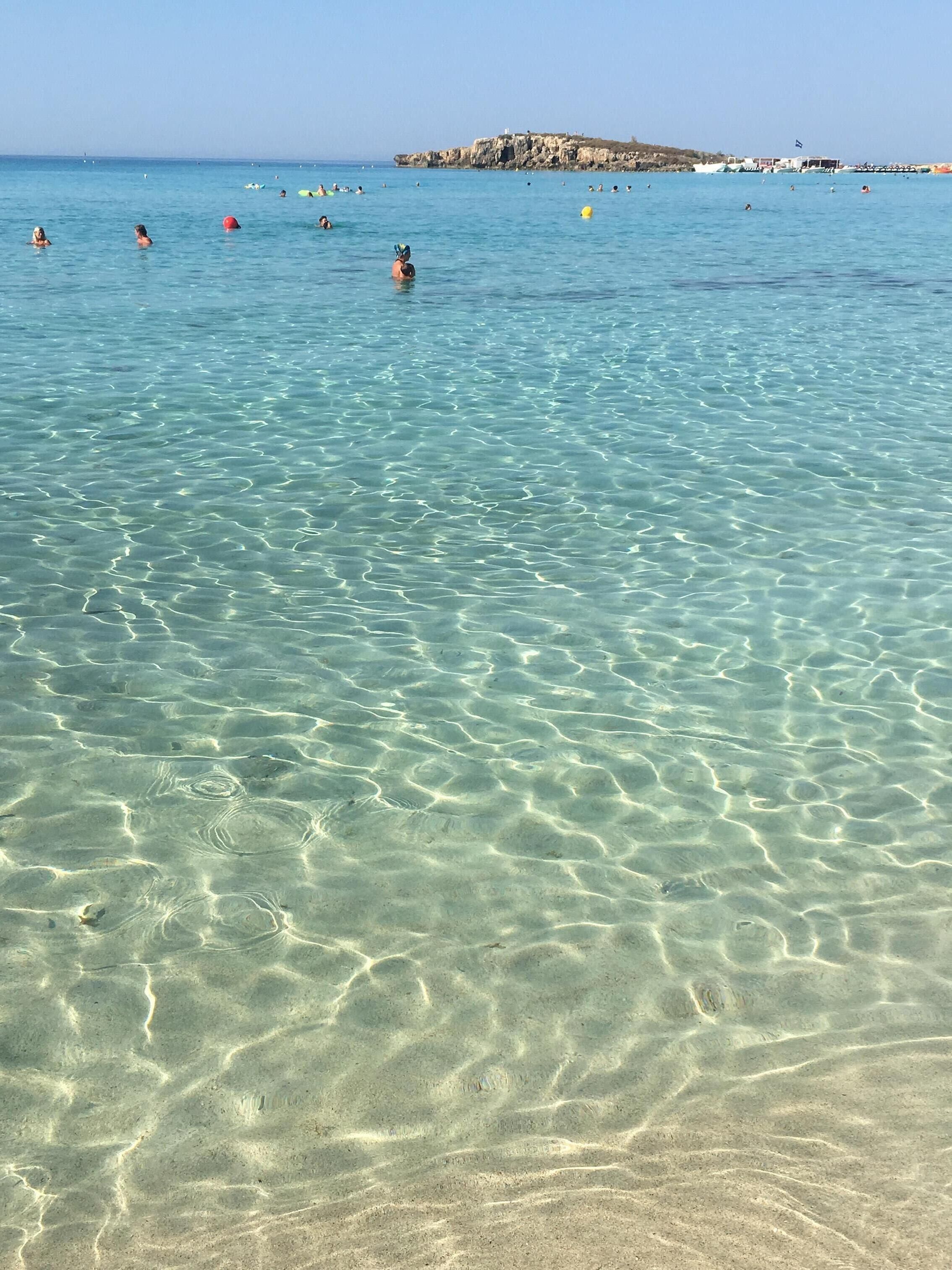 A beach with clear shallow water, people swimming, and boats in the distance.