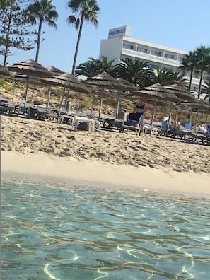A beach with clear water, umbrellas, lounge chairs, and a building labeled "Nissi Beach" in the background. - Narcissos Nissi Beach Apartment C3 (Ayia Napa)
