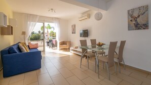 A modern living room with a blue sofa, a glass dining table with chairs, a TV, and a painting on the wall. - Narcissos Nissi Beach Apartment C3 (Ayia Napa)