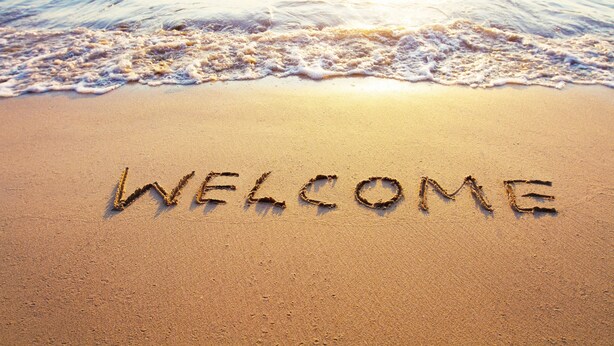 The word "WELCOME" written in the sand on a beach with waves in the background.