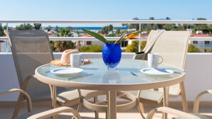 Una mesa redonda de vidrio con un florero azul y un sombrero de paja en un balcón con vistas a la costa.