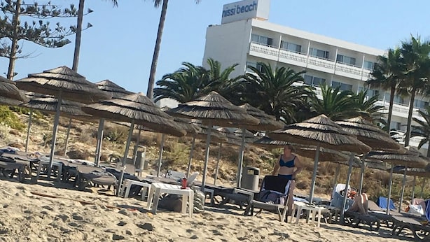 Una playa con sombrillas y sillas de descanso, un edificio con la leyenda "Nissi Beach" al fondo y agua cristalina en primer plano.