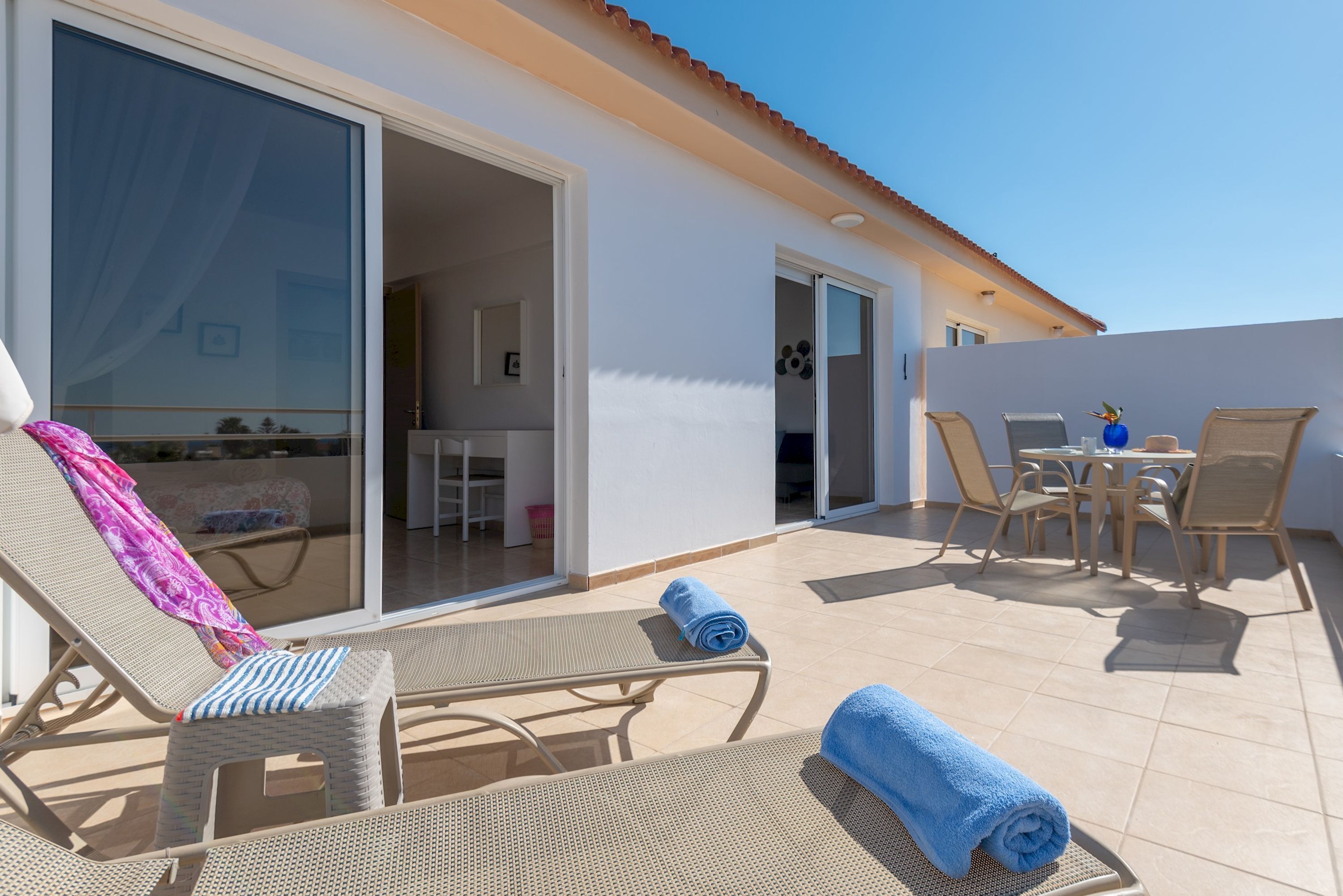 A sunlit outdoor patio with lounge chairs, a table, and a sliding glass door leading to an interior space.