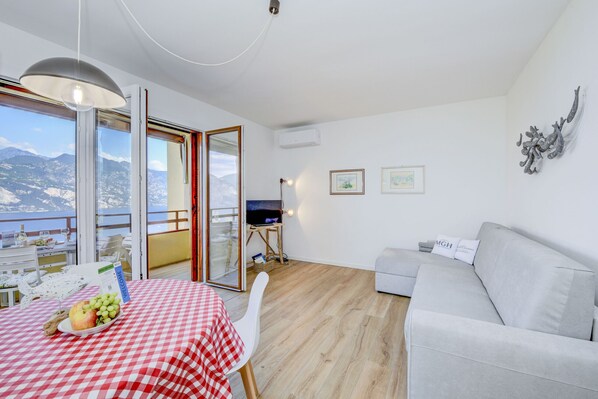 Ein modernes Wohnzimmer mit einer Couch, einem Esstisch mit kariertem Tischläufer, einem Fernseher auf einem Schrank und einem Balkon mit Blick auf die Berge.