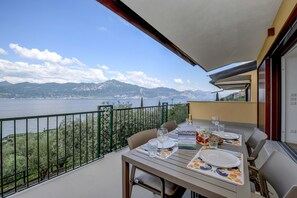 A balcony with a table set for dining, overlooking a body of water and mountains. - Pai Superior Apartment Ora (Torri del Benaco)