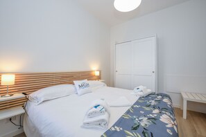 En snyggt bäddad säng med ett blommigt överkast, en sänglampa och en hopvikt handduk på nattduksbordet.