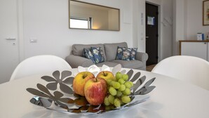 Ein Esstisch mit Obstschale, gefüllt mit Äpfeln und Trauben, ein graues Sofa mit floralen Kissen und ein großer Spiegel an der Wand.