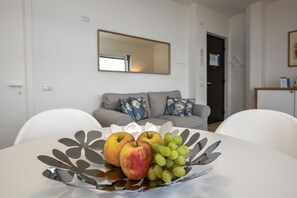 A dining table with a fruit bowl containing apples and grapes, a gray sofa with floral cushions, and a large mirror on the wall.