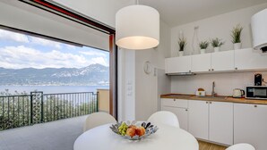 Eine moderne Küche mit Blick auf einen See und Berge, einem Esstisch mit Obstschale und weißen Schränken.