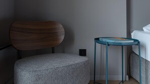 Una habitación moderna con una silla de madera, un taburete gris, una mesita de noche azul turquesa y una cama.