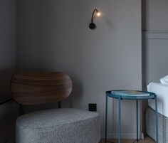 Una habitación moderna con una silla de madera, un taburete gris, una mesita de noche azul turquesa y una cama.