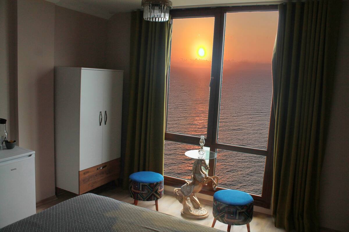 A room with a bed, a wardrobe, a small table with a lamp, two chairs, and a view of the sunset over the water.