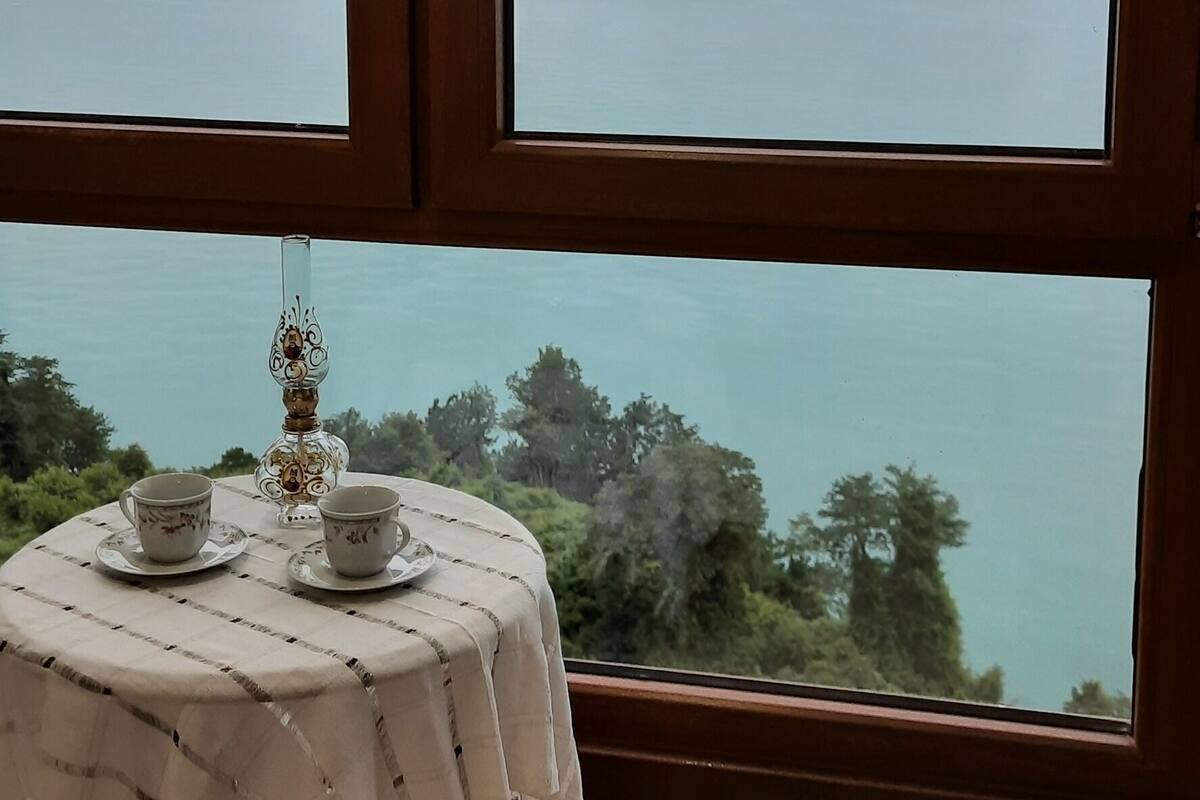A room with a large window overlooking a body of water and a sunset. There is a table with two cups and saucers, and a decorative candle holder.