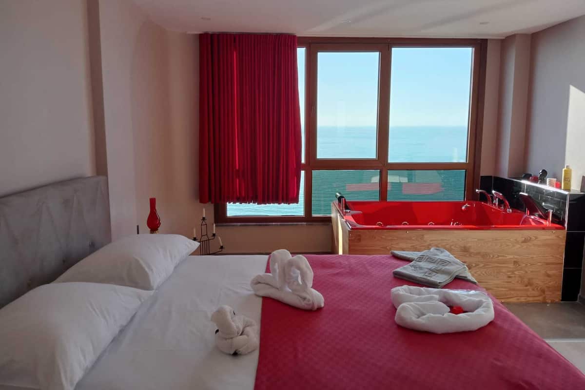 A hotel room with a bed, a red bath tub, and a large window with a view of the sea.