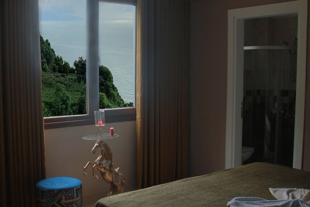 A hotel room with a large bed, a small table with a chair, a window with a view of the sea, and a door leading to a bathroom.