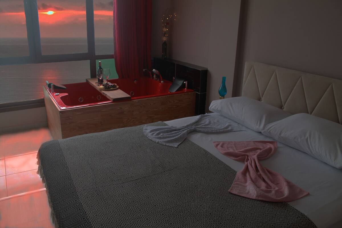 A bedroom with a bed, a nightstand, a red sink, and a view of the sunset through large windows.