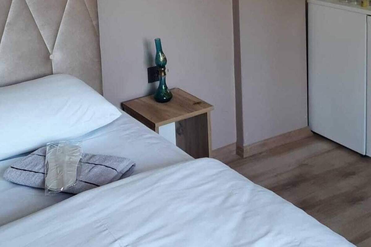 A hotel room with a bed, a nightstand with a green vase, a white cabinet, and a wooden floor.