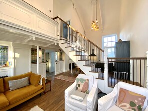 A spacious living area with a staircase, a yellow sofa, white armchairs, and a kitchen in the background. - The Chapel Hayle (Hayle)