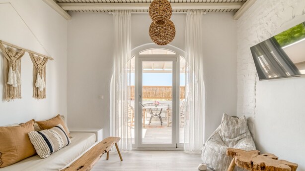 Un salón luminoso y espacioso con un banco de madera, un sofá con cojines estampados, una lámpara de techo grande de fibra natural, y vista del patio exterior a través de un ventanal.