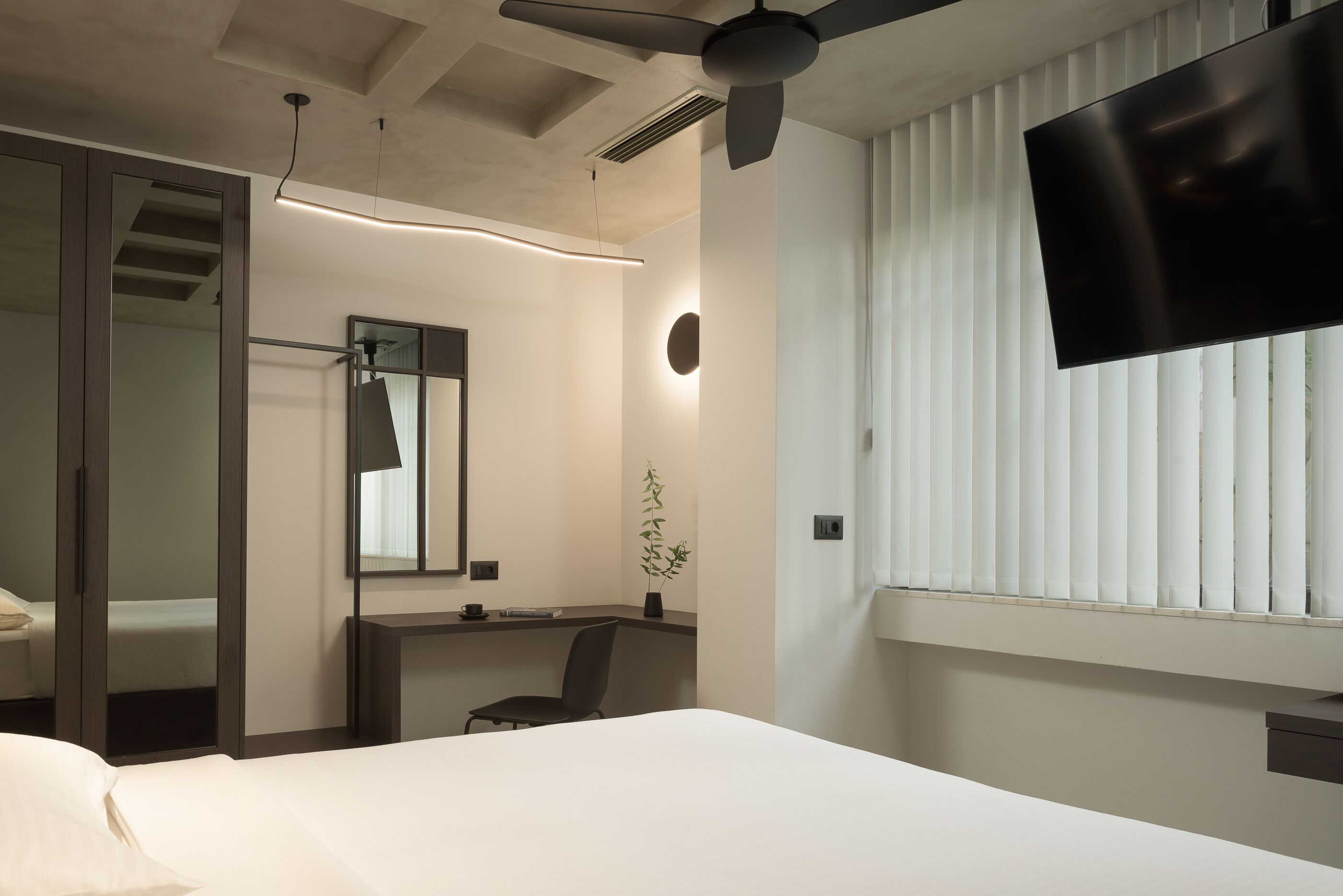 Hotelkamer met een bed, bureau, stoel, spiegel, plafondventilator, televisie en raam met zonwering.