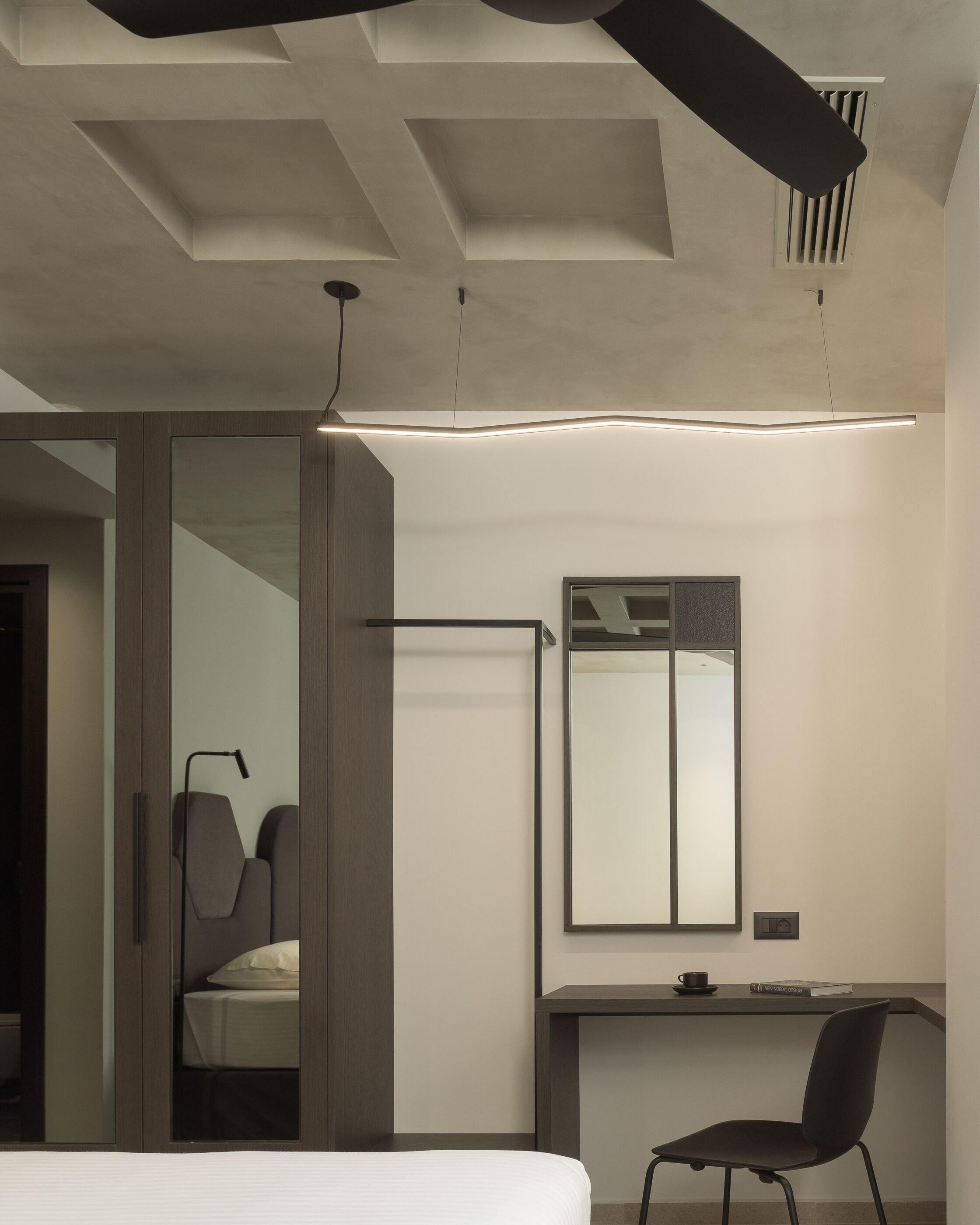 A modern hotel room with a bed, a desk, a chair, a large mirror, and a ceiling light fixture.