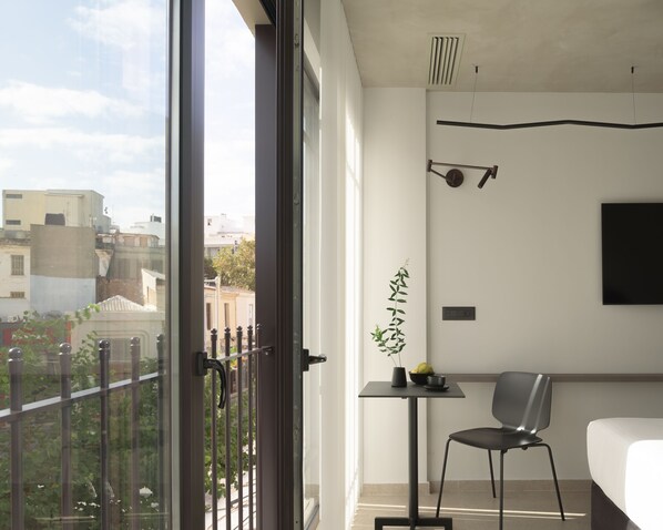 A modern hotel room with a large balcony, a bed, a chair, a small table with a plant, and a wall-mounted TV. - Pleiades Urban Stay by Semavi (Heraklion)