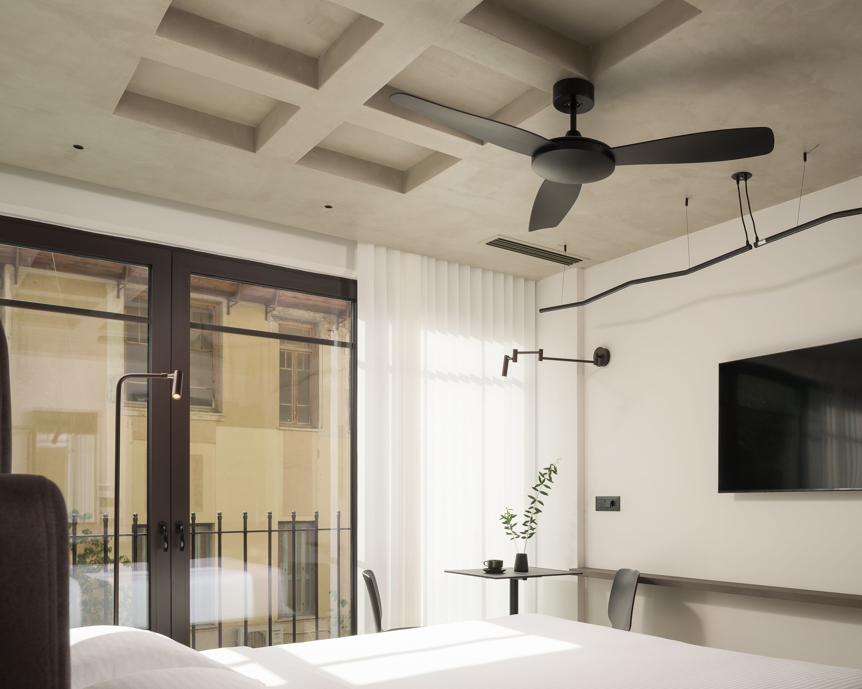 A hotel room with a bed, a desk, a chair, a TV mounted on the wall, a ceiling fan, and a view of the outside through a sliding glass door.