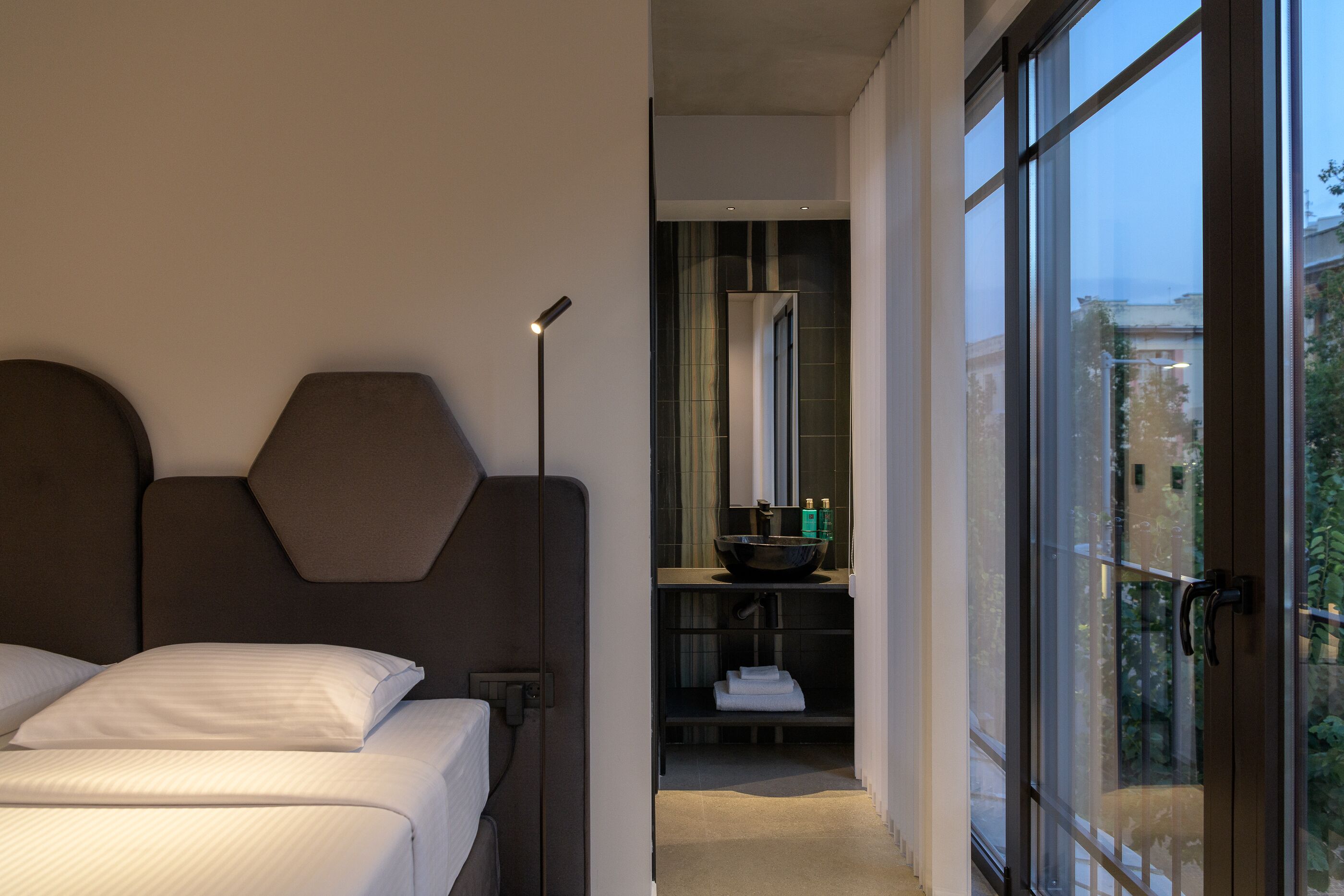 A modern hotel room with a large bed, a nightstand, a bedside lamp, a bathroom with a large mirror, and a view of the city through the window.