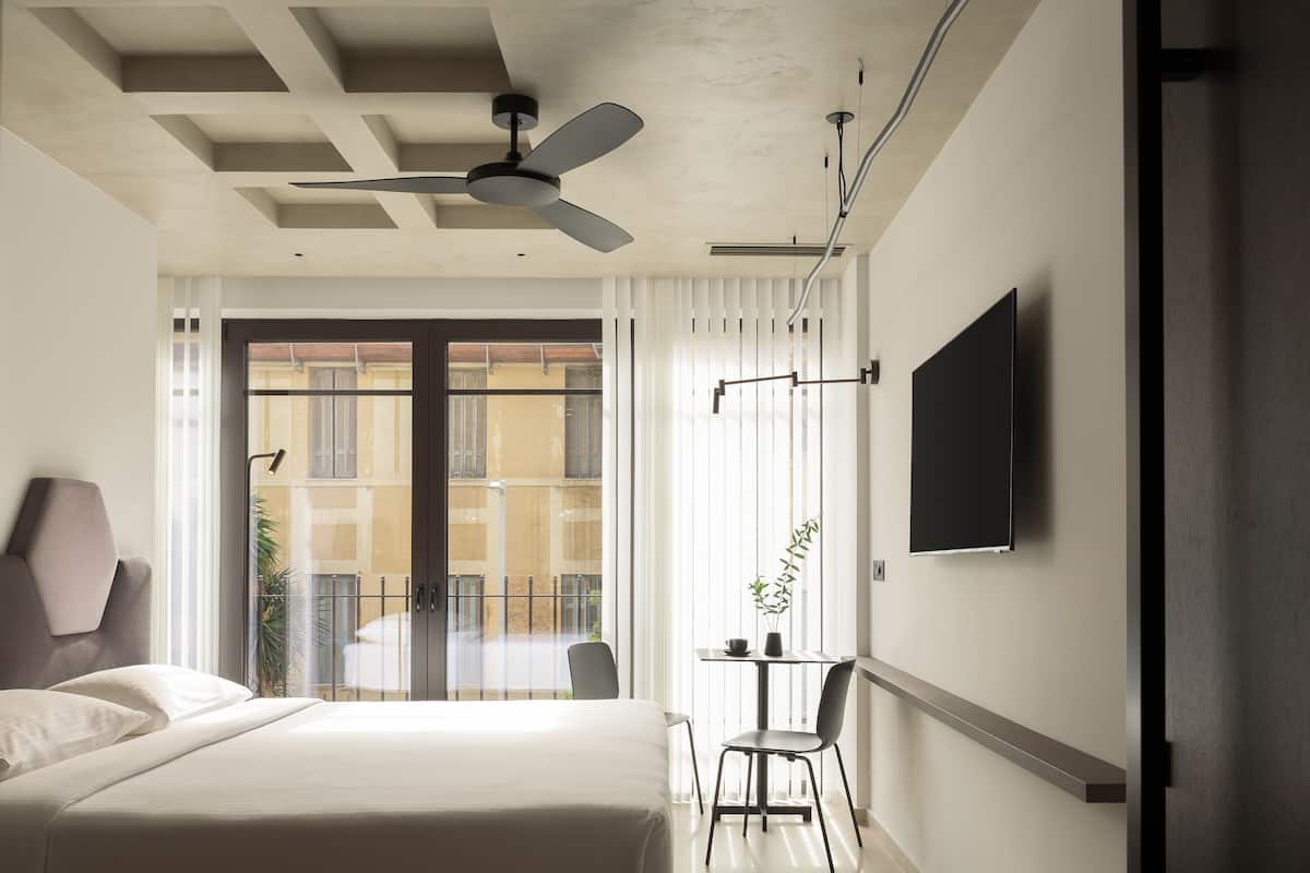 A modern hotel room with a large bed, a ceiling fan, a TV mounted on the wall, and a balcony with a view of the city.