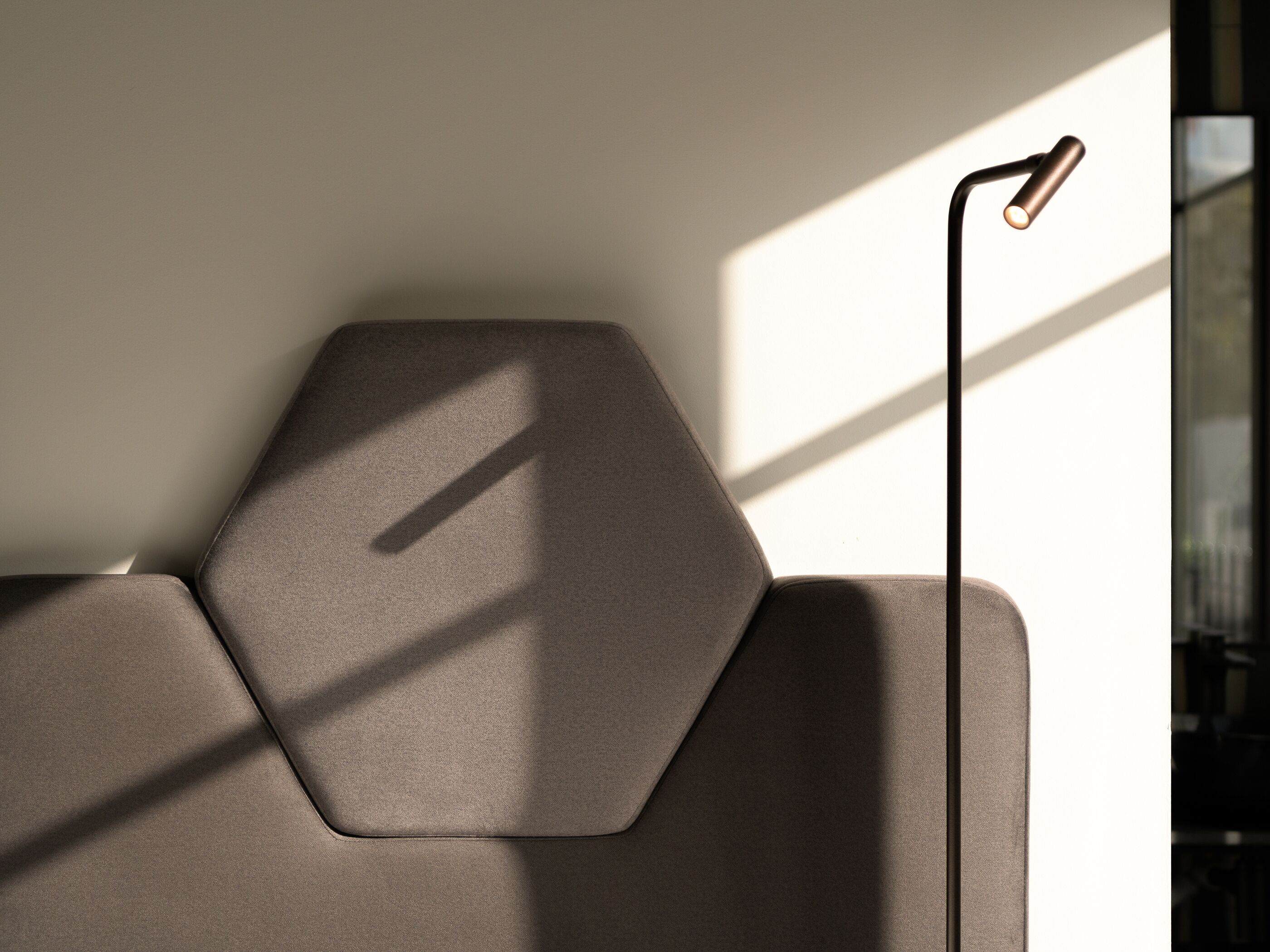 Un canapé moderne et minimaliste, doté d’un design géométrique, est placé contre un mur percé d’une grande fenêtre, ce qui permet à la lumière naturelle de créer des motifs sur la surface.