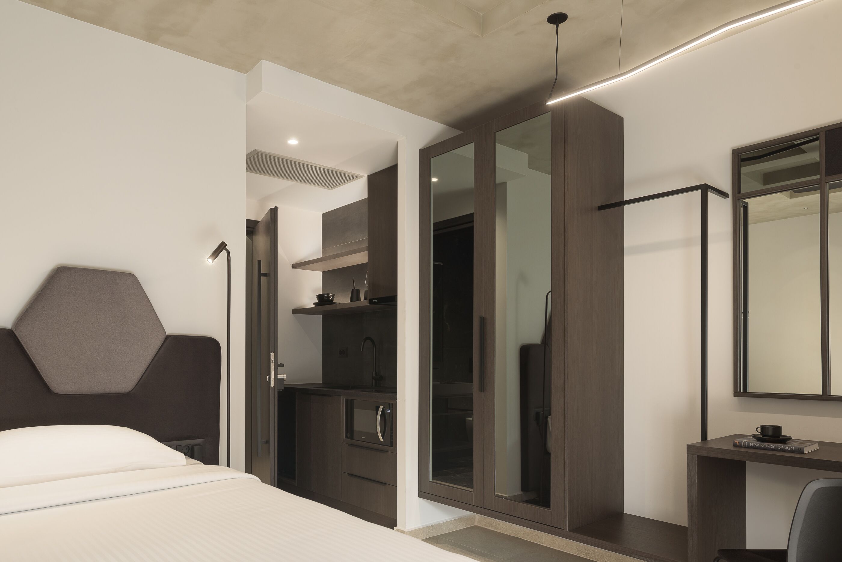 A modern hotel room with a large bed, a desk, a mirror, and a kitchenette.