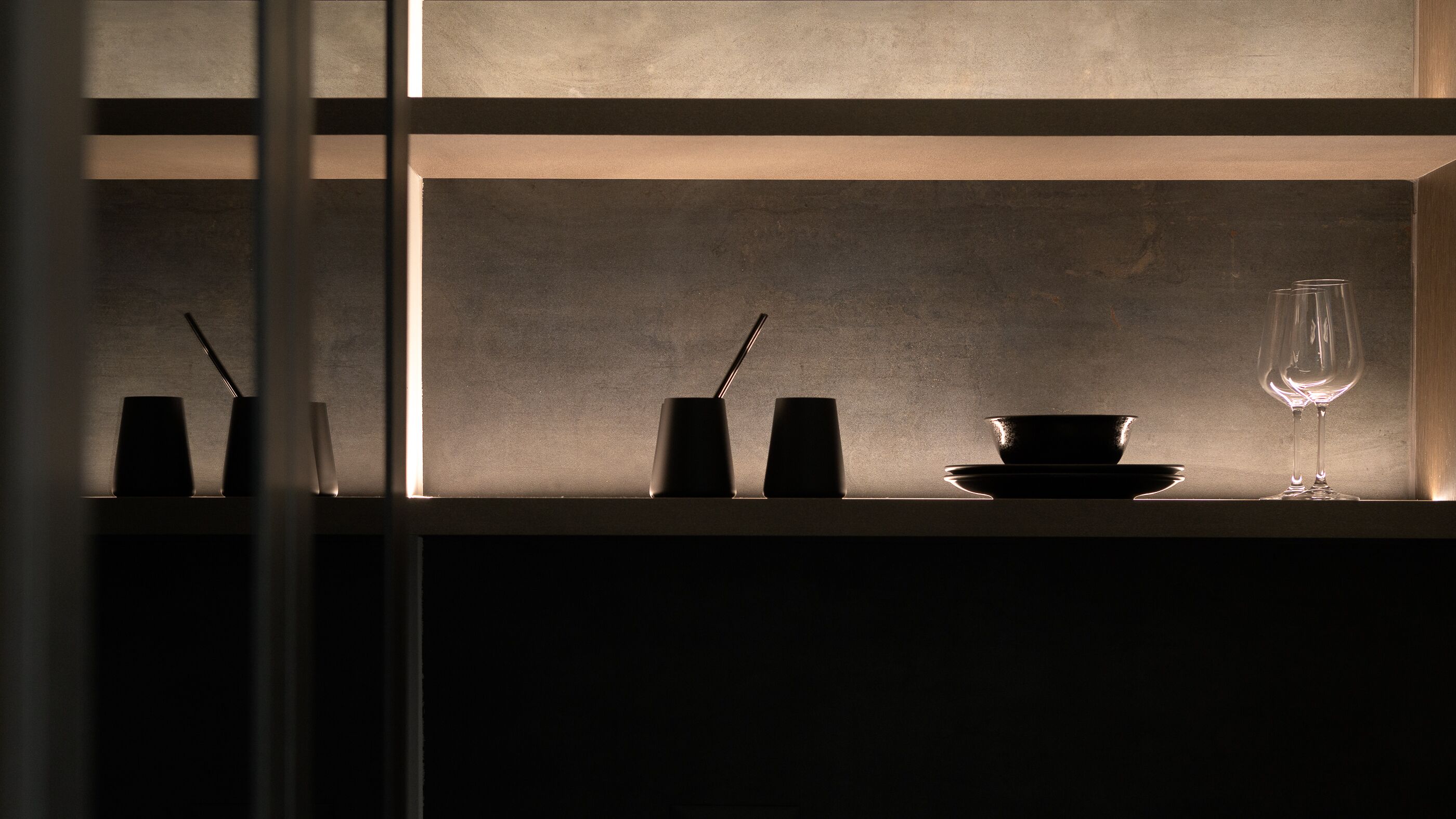 A dimly lit interior with a shelf displaying a black bowl, a wine glass, and two black candle holders.