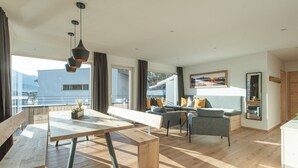 A modern living room with a wooden floor, a dining area with a bench and table, a grey sofa with cushions, and a large window offering a view of the outdoors. - Apartment Auszeitmit in the Salzburger Sportwelt vacation region (Eben im Pongau)