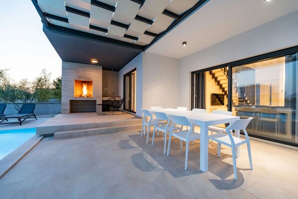 A modern outdoor patio with a pool, a dining area with white chairs, and a fireplace.